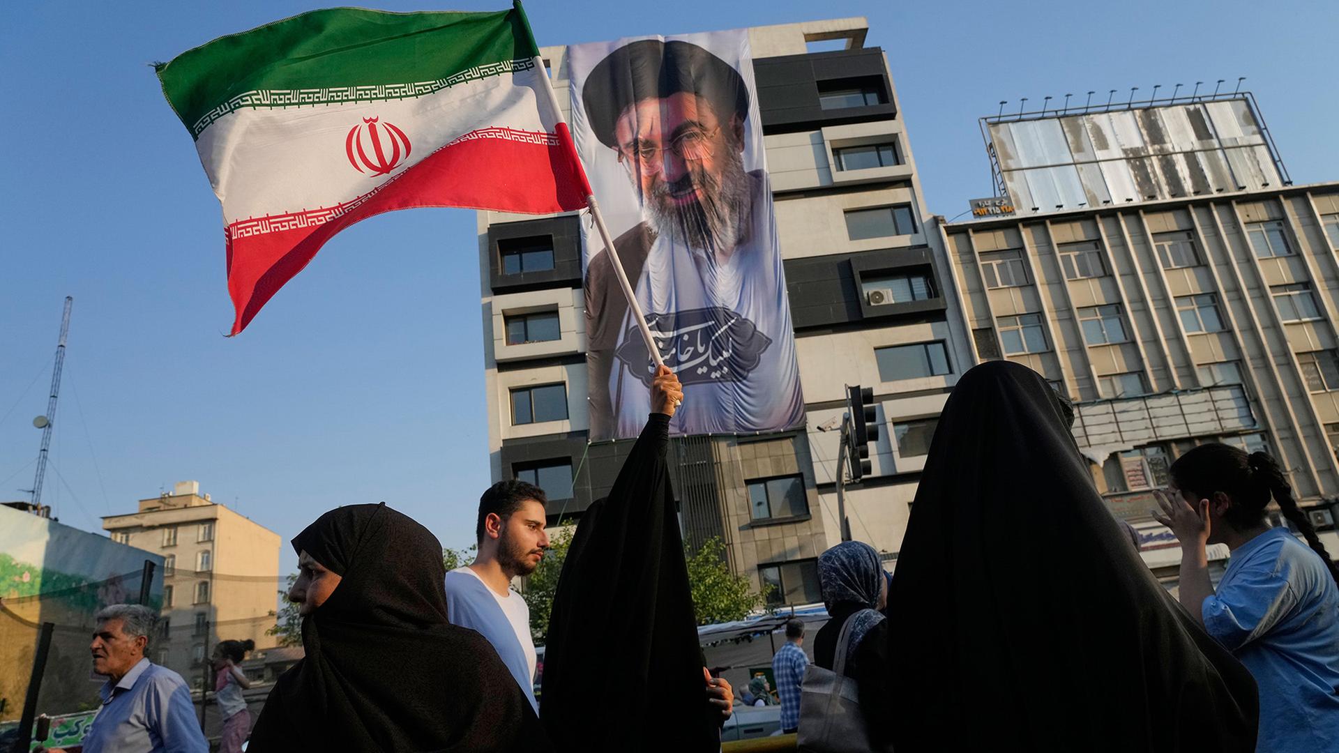 Eine Frau hält eine iranische Flagge vor einem Banner mit einem Porträt des Obersten Führers Ayatollah Mojtaba Khamenei während eines Straßenfestes. | Vahid Salemi/AP/dpa