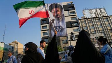 Eine Frau hält eine iranische Flagge vor einem Banner mit einem Porträt des Obersten Führers Ayatollah Mojtaba Khamenei während eines Straßenfestes.