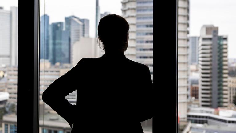 Silhouette einer Frau, die in einem Büro im oberen Stock eines Hochauses aus dem Fenster schaut.