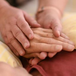 Eine Hospizmitarbeiterin hält die Hand eines todkranken Menschen, der im Hospiz im Bett liegt.