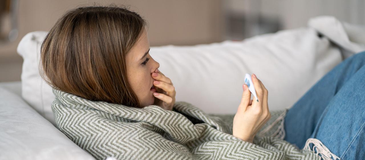 Eine hustende Frau schaut auf ein Fieberthermometer.