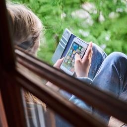 Eine jugendliche Person sitzt am Fenster und schaut auf sein Smartphone.