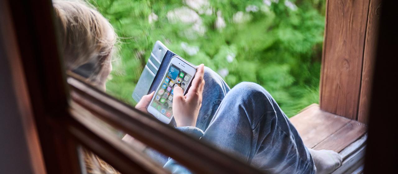 Eine jugendliche Person sitzt am Fenster und schaut auf ihr Smartphone.