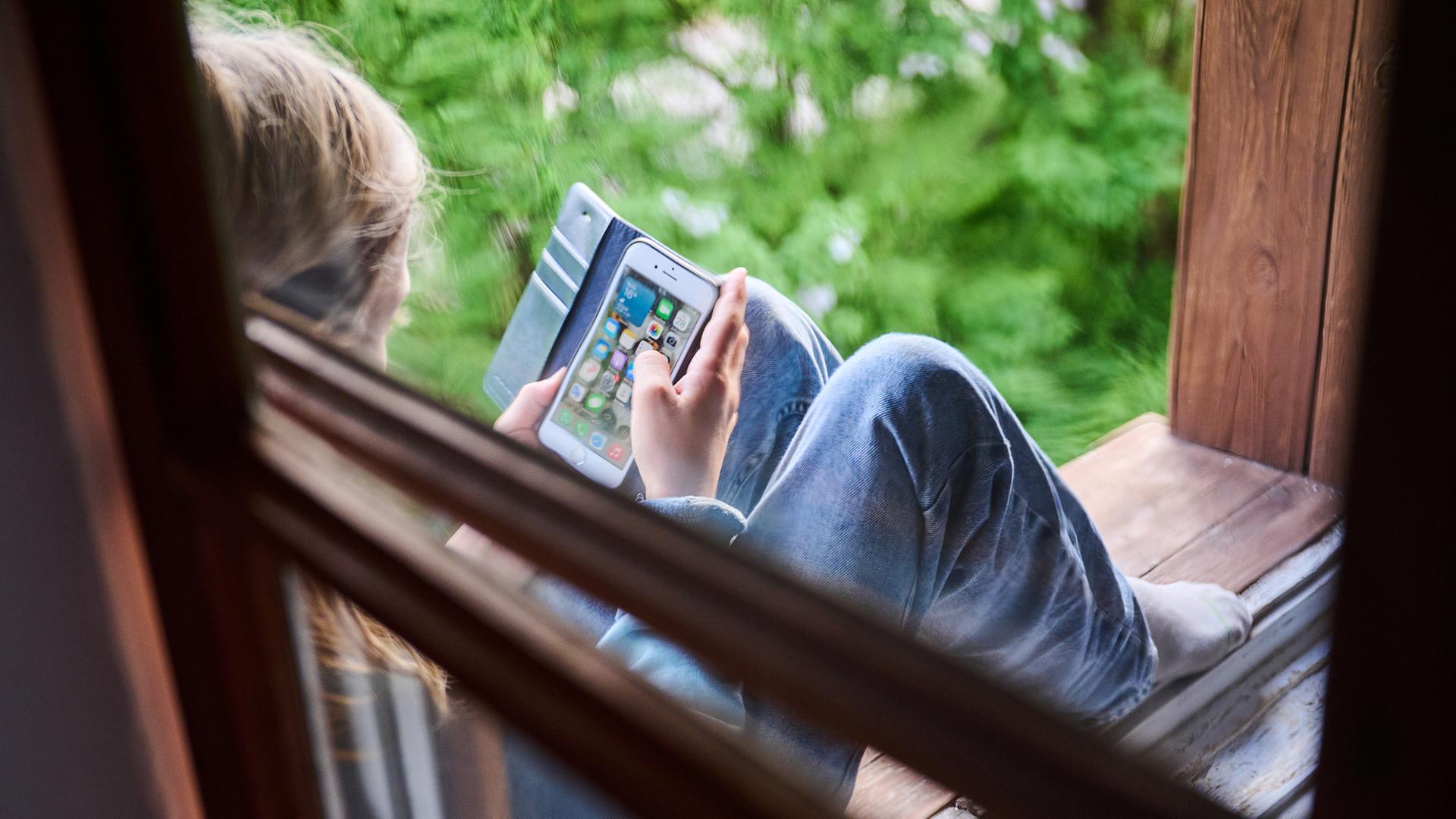 Eine jugendliche Person sitzt am Fenster und schaut auf ihr Smartphone. | picture alliance/dpa