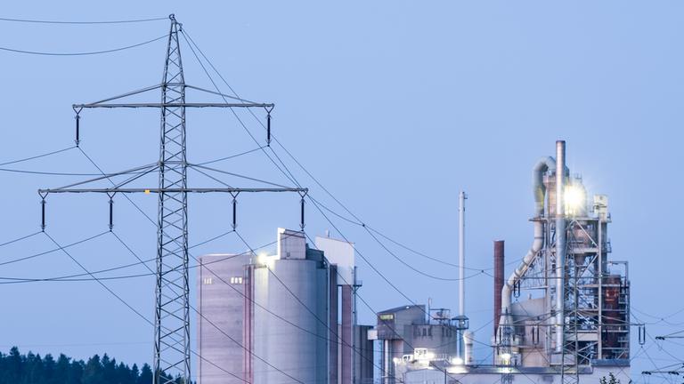 Strommasten stehen im Morgenlicht vor einem Zementwerk.