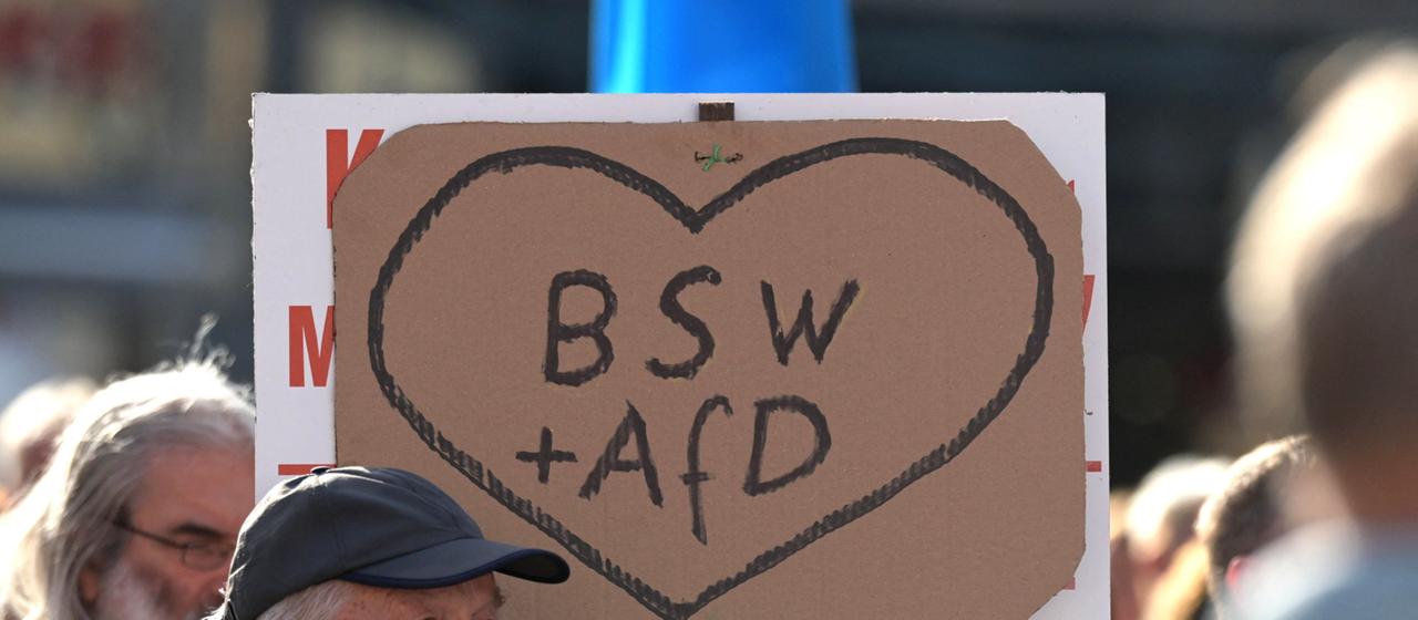 Ein Schild mit einem Herz und der Aufschrift BSW + AfD wird von Teilnehmern einer Wahlveranstaltung hochgehalten.