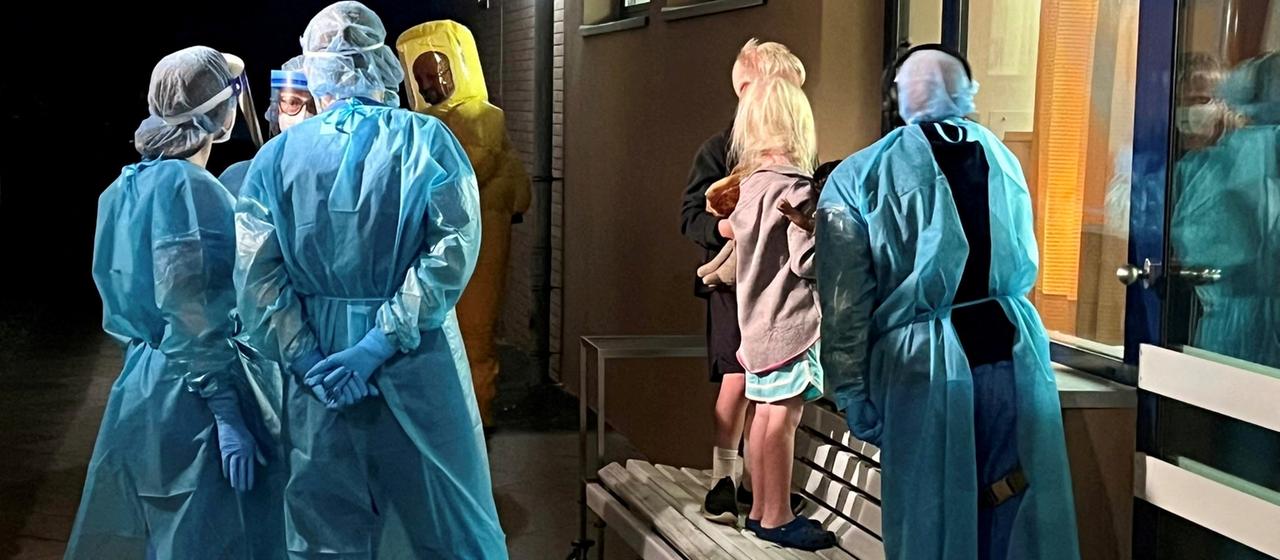 Medizinisches Personal in Schutzkleidung und zwei Kinder schauen durch eine Glasscheibe