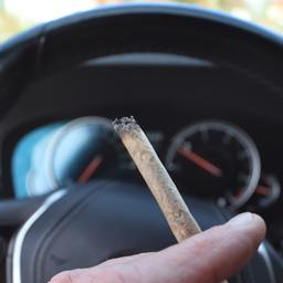 Ein Mann sitzt mit einem Joint zwischen den Fingern am Steuer eines Autos.