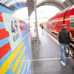 Ein Mann geht an einem Bahnhof mit seinem Fahrrad zwischen der Werbung für das Deutschlandticket und einer Regionalbahn entlang. 