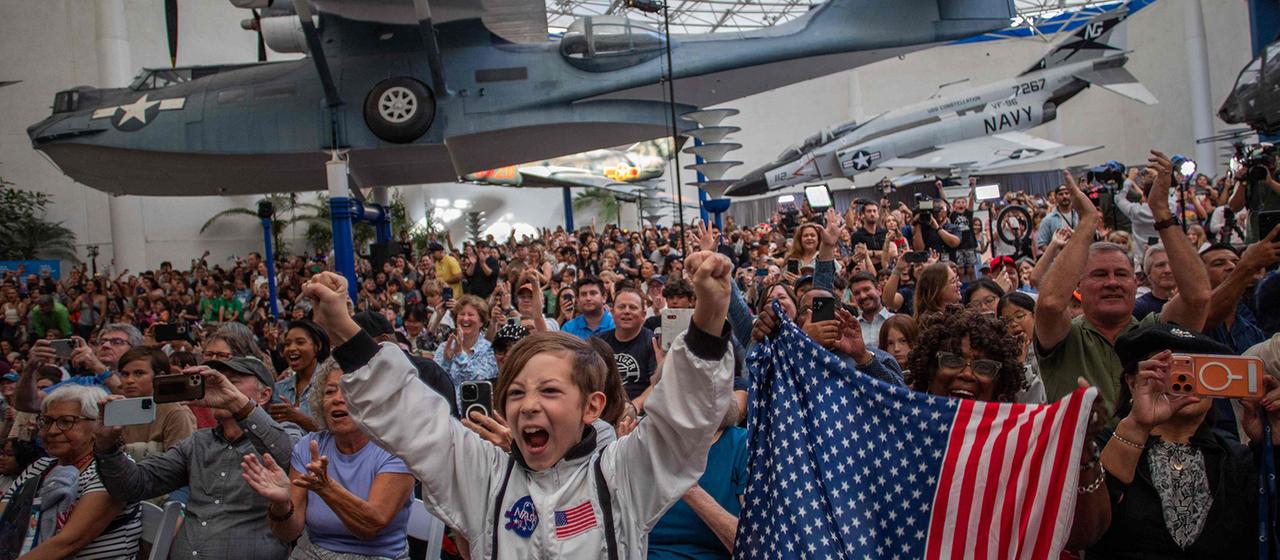 Menschen jubeln in einer Halle, in der historische Militärflugzeuge ausgestellt sind.