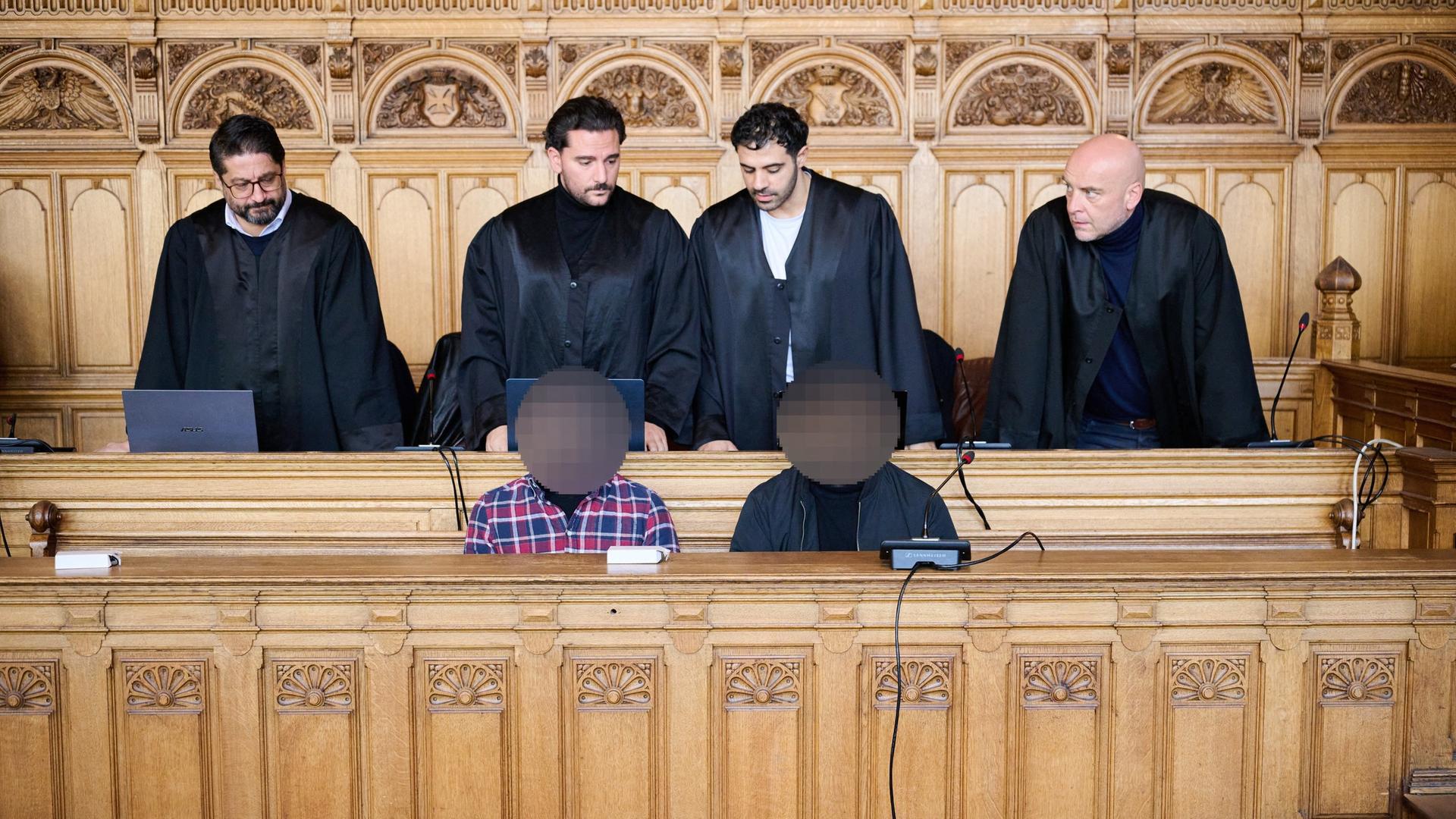 Zwei Männer, deren Gesichter unkenntlich gemacht wurden, sitzen auf der Angeklagebank eines Gerichtes. Hinter ihnen stehen vier Männer in Roben. | dpa | Sina Schuldt