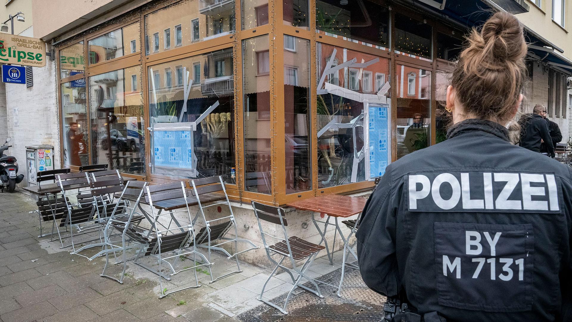 Eine Polizistin steht vor einem israelischen Restaurant in München, in dem nach einem Angriff zerbrochene Scheibe notdürftig repariert wurden. | Peter Kneffel/dpa