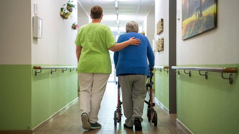 Eine Pflegefachkraft geht mit einer Seniorin den Flur einer Pflegeeinrichtung entlang.