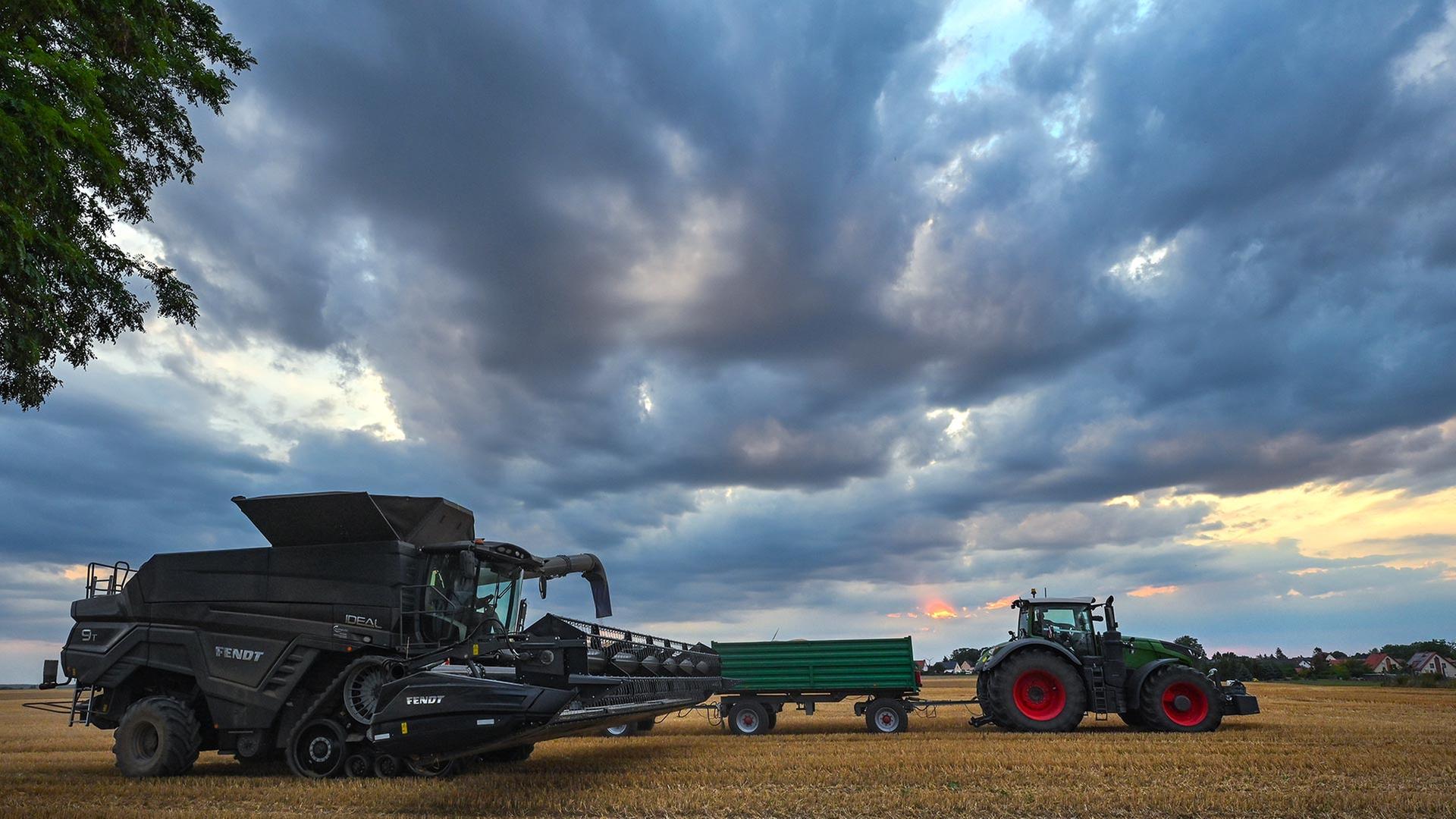 Ein Mähdrescher und ein Traktor mit Anhänger stehen im Sonnenuntergang auf einem Feld in Ostbrandenburg.