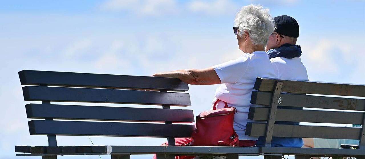 Eine ältere Frau und ein älterer Mann sitzen auf einer Bank in der Sonne.