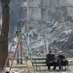 Zwei Männer sitzen auf einer Spielplatz-Bank vor zerstörten Häusern in Mariupol.