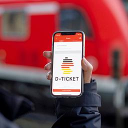 Eine Frau hält ihr Smartphone hoch, sodass das Logo des Deutschlandtickets deutlich auf ihrem Smartphone zu erkennen ist - im Hintergund befindet sich ein roter Zug.