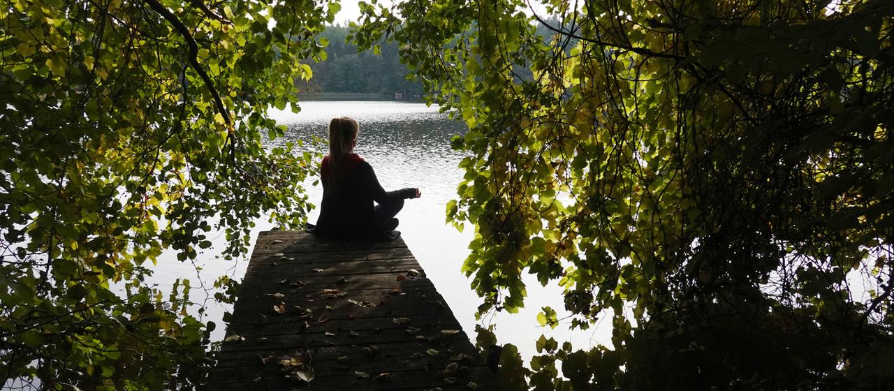  Frau sitzt allein auf einem Steg an einem See und meditiert.