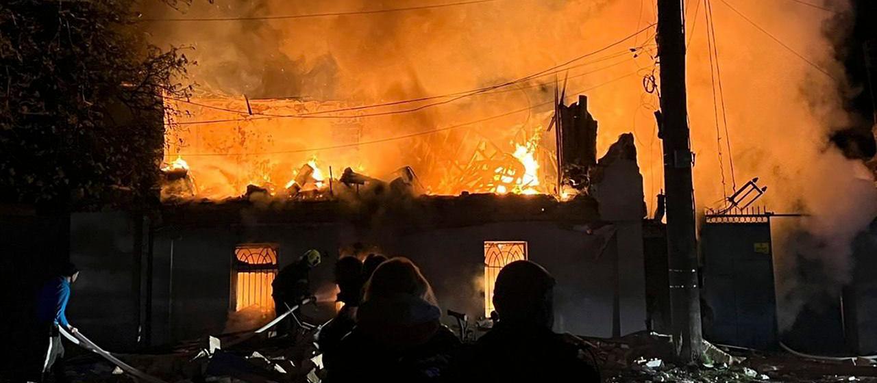 Menschen stehen vor einem zerstörten, brennenden Haus.