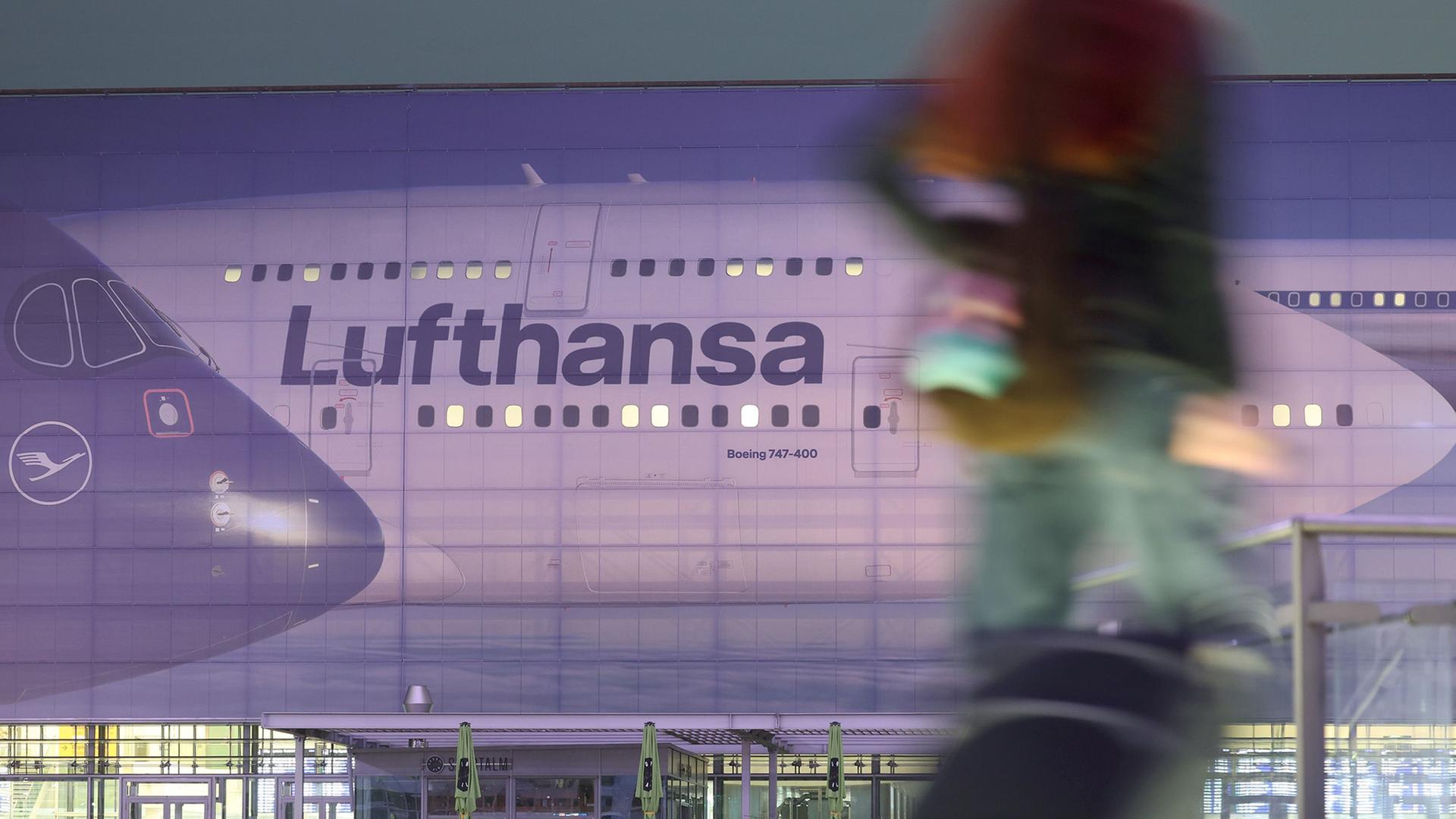 Menschen gehen am frÃ¼hen Morgen am Terminal 2 vor einer groÃen Abbildung einer Lufthansa-Maschine. | Karl-Josef Hildenbrand/dpa