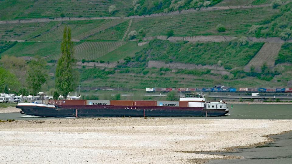 Ein mit bunten Containern beladenes Frachtschiff auf dem Wasser, daneben eine Sandbank.