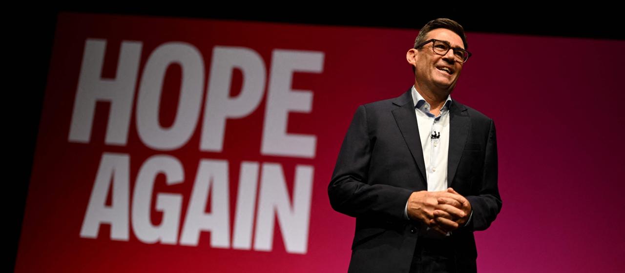 Andy Burnham steht vor einem roten Hintergrund, auf dem "Hope again" steht.
