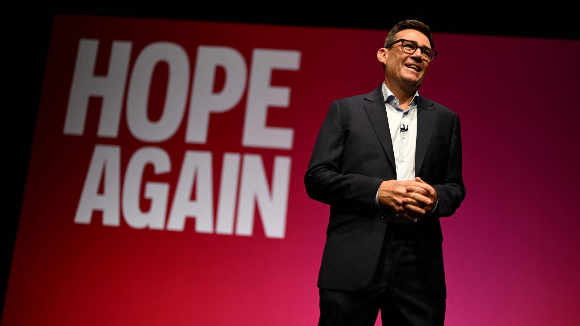 Andy Burnham steht vor einem roten Hintergrund, auf dem 