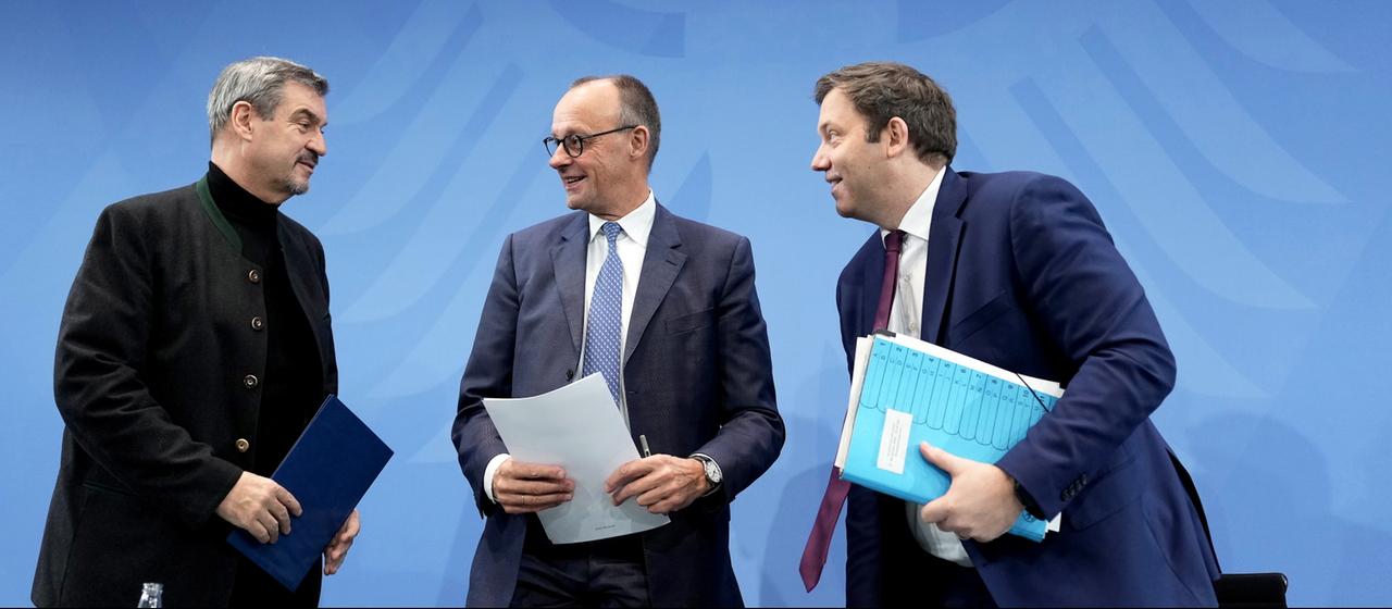 Friedrich Merz steht zwischen Lars Klingbeil und Markus Söder. Alle lächeln und halten Papiere in der Hand.