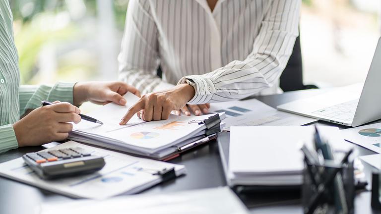 Zwei Menschen in einer Besprechung an einem Schreibtisch auf dem sich Dokumente, ein Laptop und ein Taschenrechner befinden.