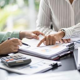 Zwei Menschen in einer Besprechung an einem Schreibtisch auf dem sich Dokumente, ein Laptop und ein Taschenrechner befinden.