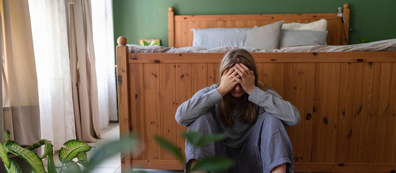 Eine Frau mit den Händen vor dem Gesicht sitzt vor einem Bett.