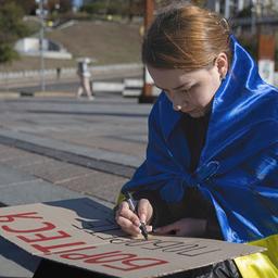Eine junge Frau mit ukrainischer Flagge um die Schultern schreibt auf ein Pappschild.