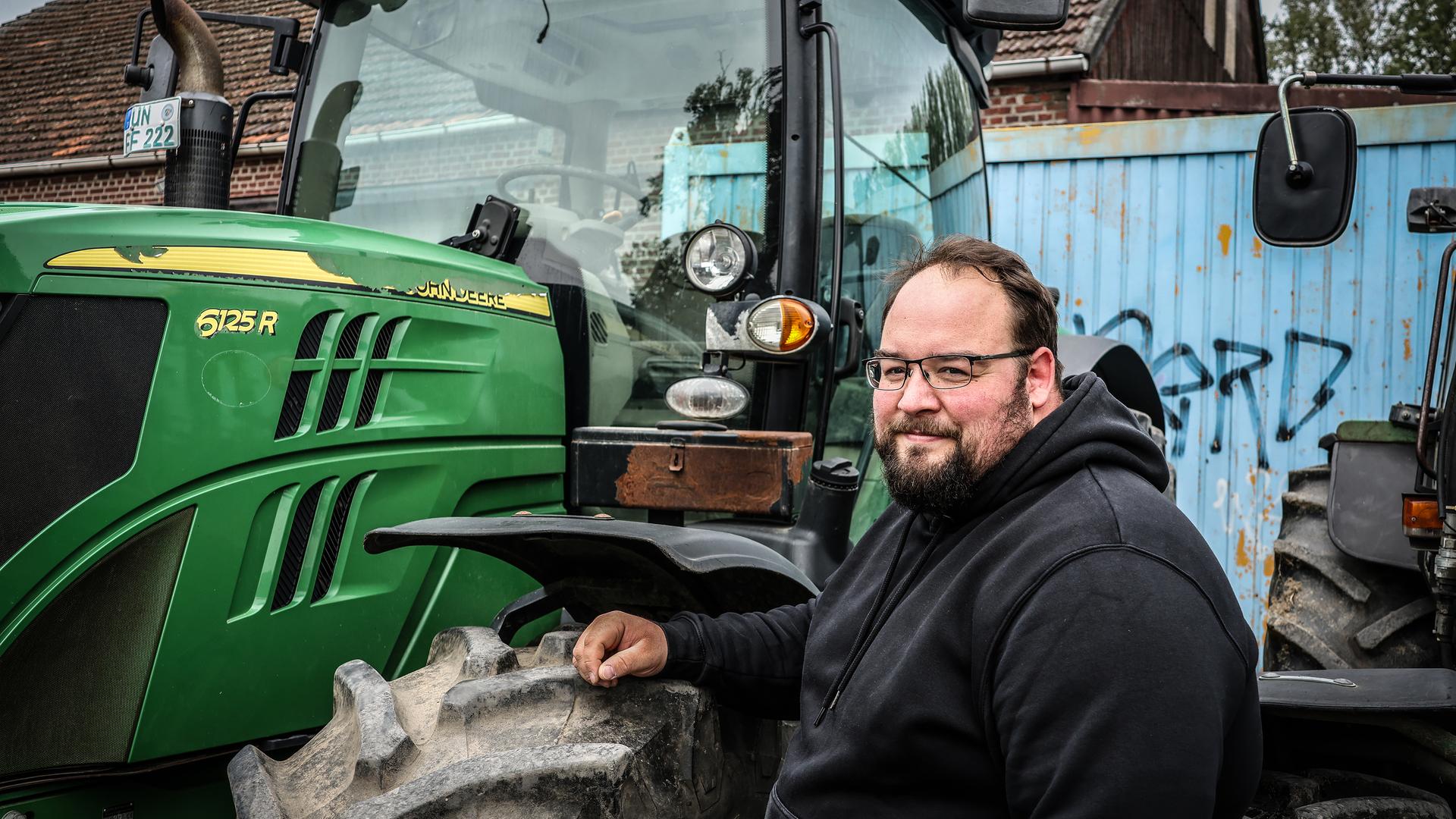 Landwirt Julian Freisendorf steht im schwarzen Pullover neben seinem grünen Traktor. | WDR / Dirk Planert