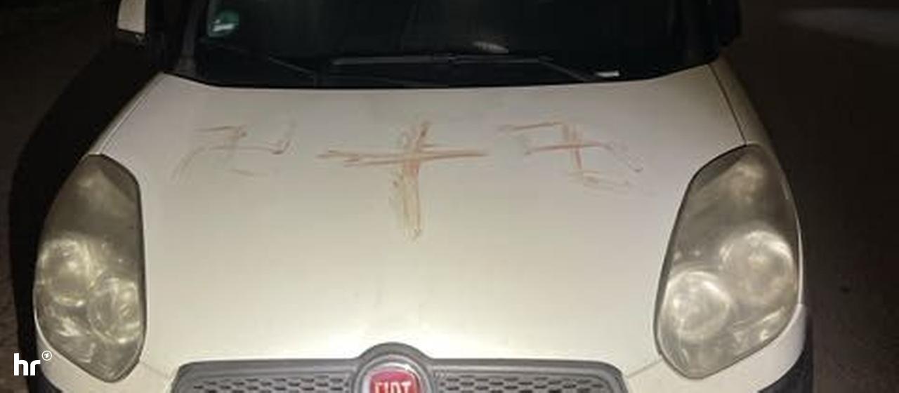 Nachts sind auf der Motorhaube eines weißen Autos drei blutverschmierte (Haken-)Kreuze zu sehen