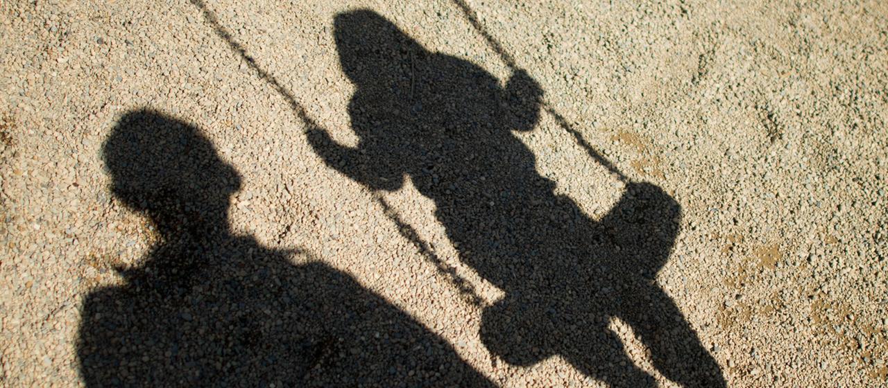 Der Schatten von einem Mann und einem schaukelnden Kind fallen auf Sand auf einem Spielplatz. 