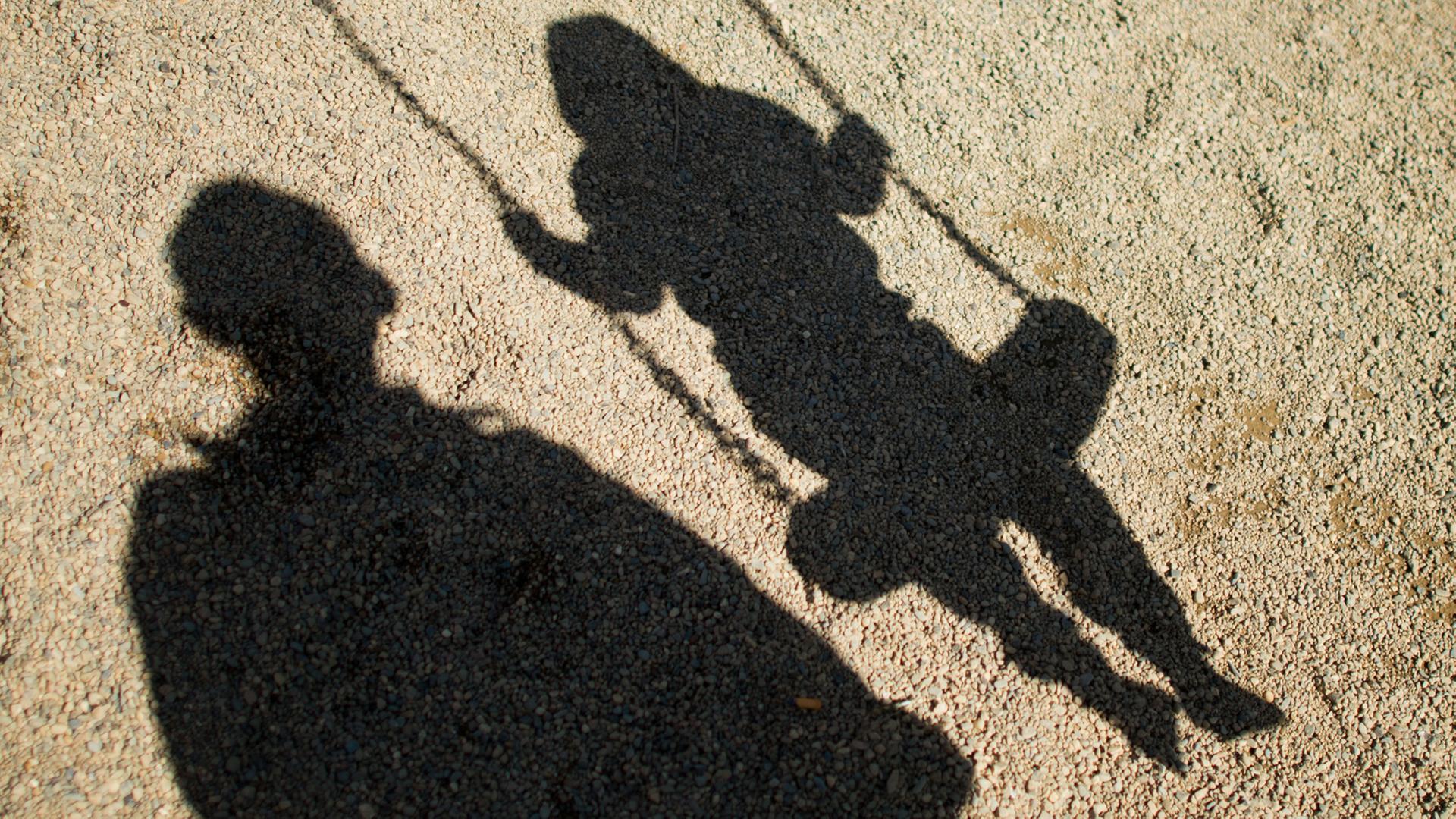 Der Schatten von einem Mann und einem schaukelnden Kind fallen auf Sand auf einem Spielplatz.  | dpa