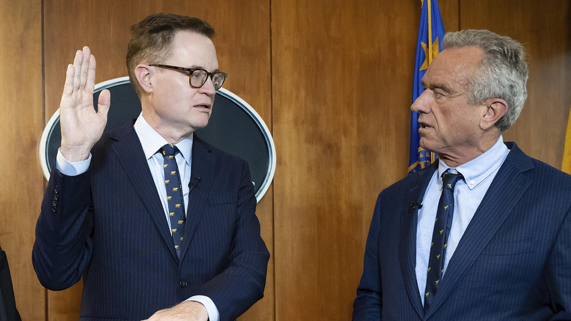 Jim O'Neill und Robert Kennedy Junior stehen im Ministerium. O'Neill hält die Hand hoch zum Schwur.  | AP