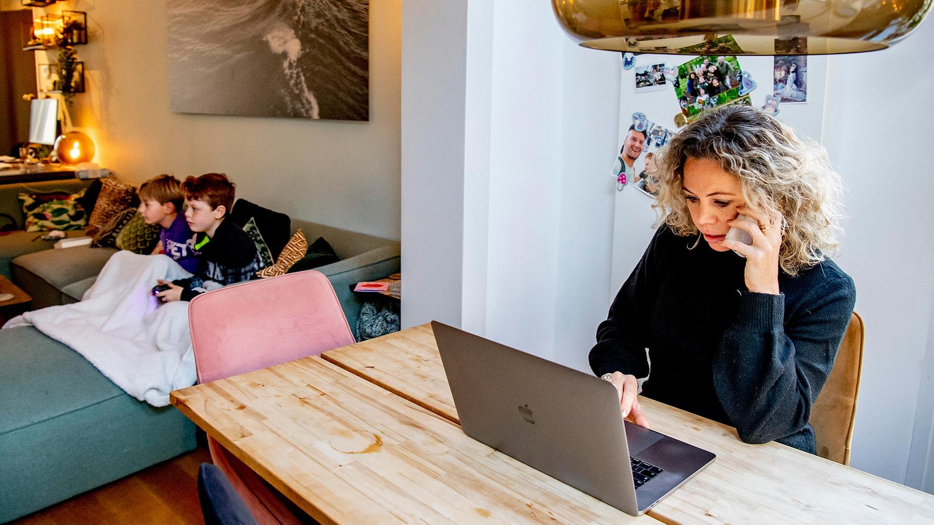 Eine Frau mit einem Laptop am Esszimmertisch, Kinder sitzen im Hintergrund. | picture alliance / ROBIN UTRECHT