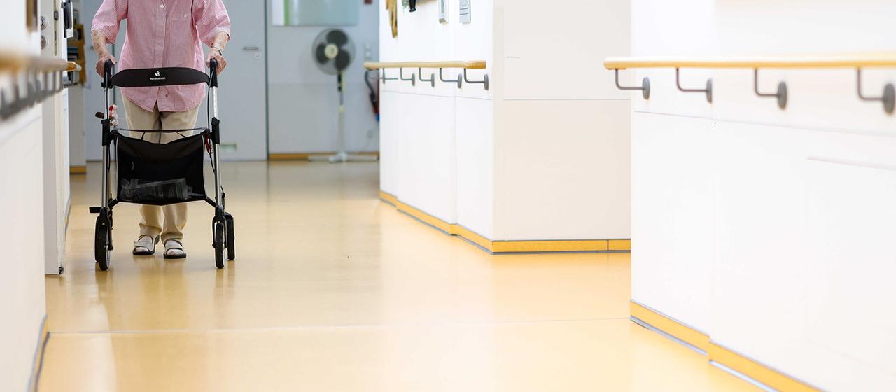 Eine ältere Frau geht mit einem Rollator über einen Flur.