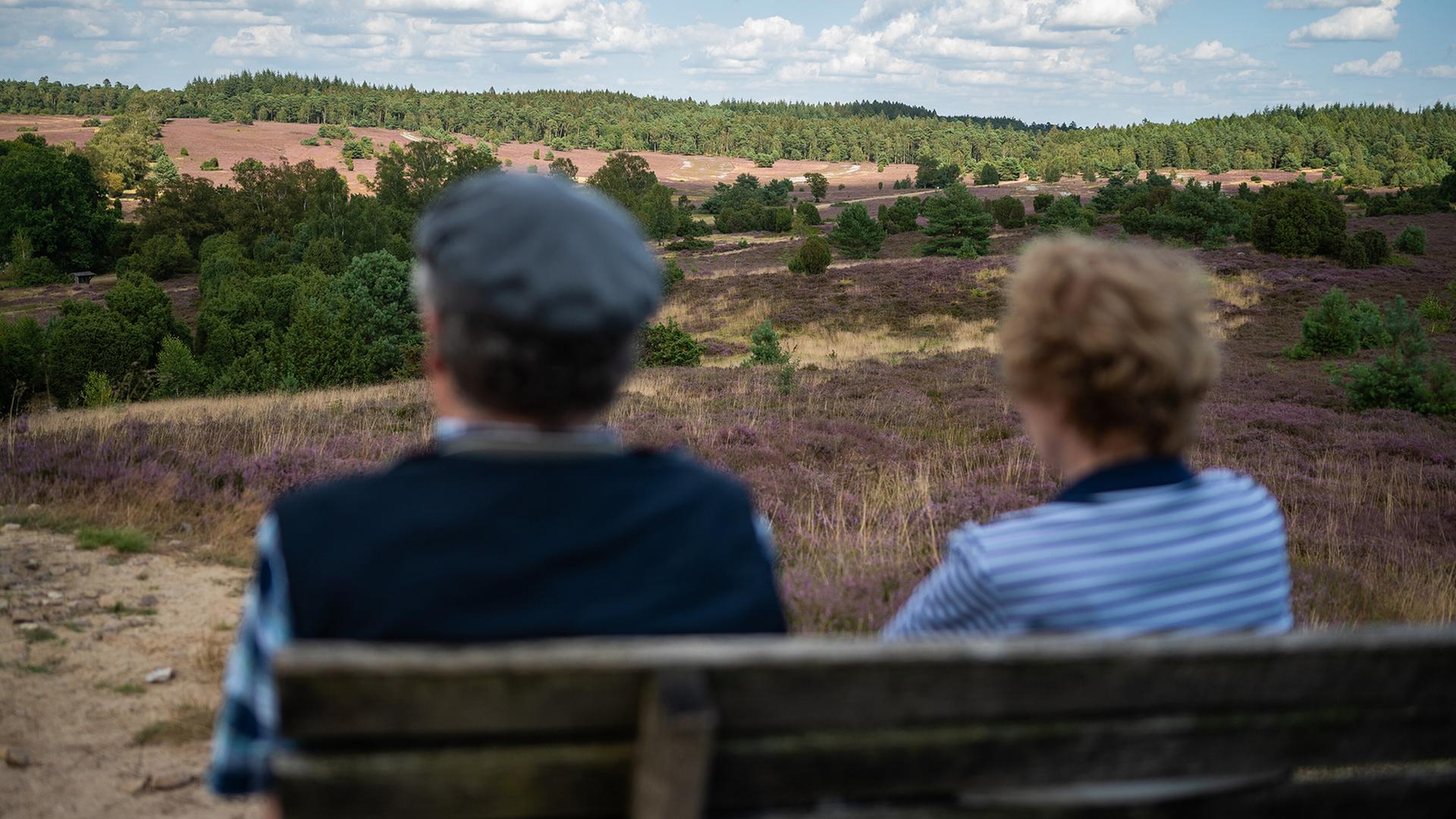 Zwei Menschen sitzen auf einer Bank in der Heidelandschaft. | picture alliance/dpa