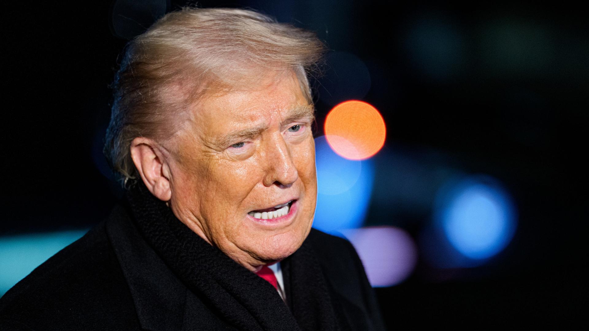 Donald Trump in einem schwarzen Mantel, im Hintergrund verschwommen Signallichter de Flughafens in Washington. | EPA