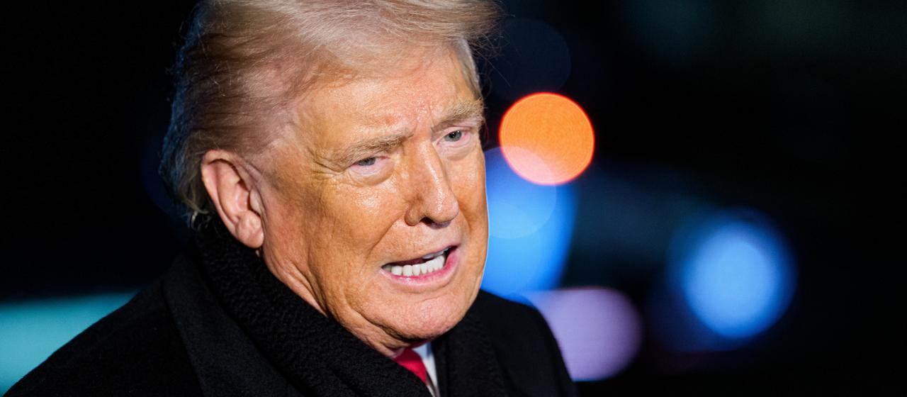 Donald Trump in einem schwarzen Mantel, im Hintergrund verschwommen Signallichter de Flughafens in Washington.