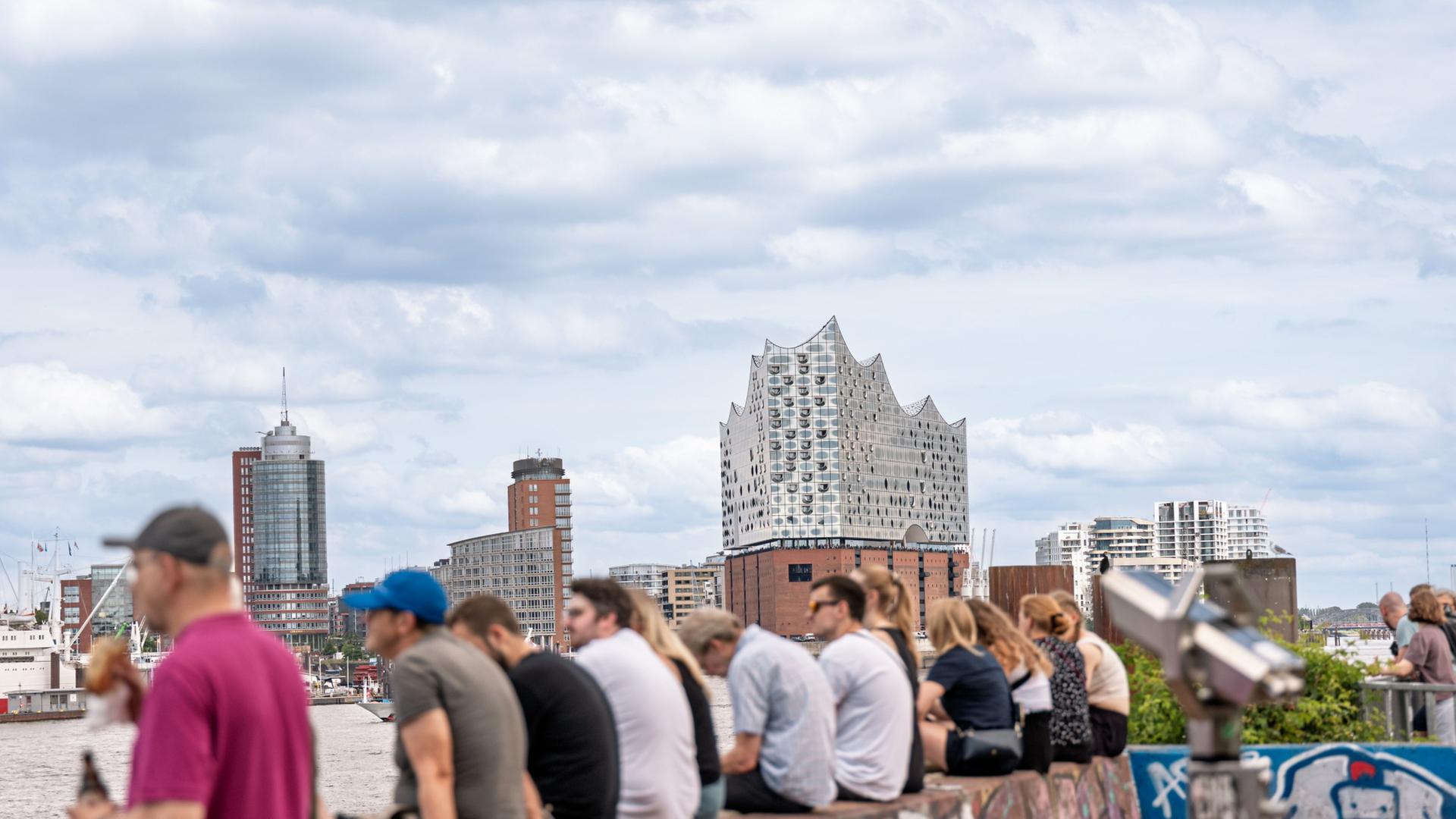 Menschen sitzen in Hamburg auf einer Mauer mit Blick auf die Elbphilharmonie. | Markus Scholz/dpa
