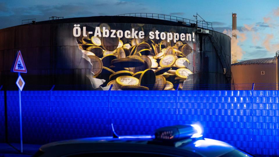 Greenpeace-Aktivisten projizieren den Schriftzug "Öl-Abzocke stoppen" auf einen großen Tank einer Raffinerie im Hamburger Hafen, im Vordergrund Blaulicht eines Polizeiautos.