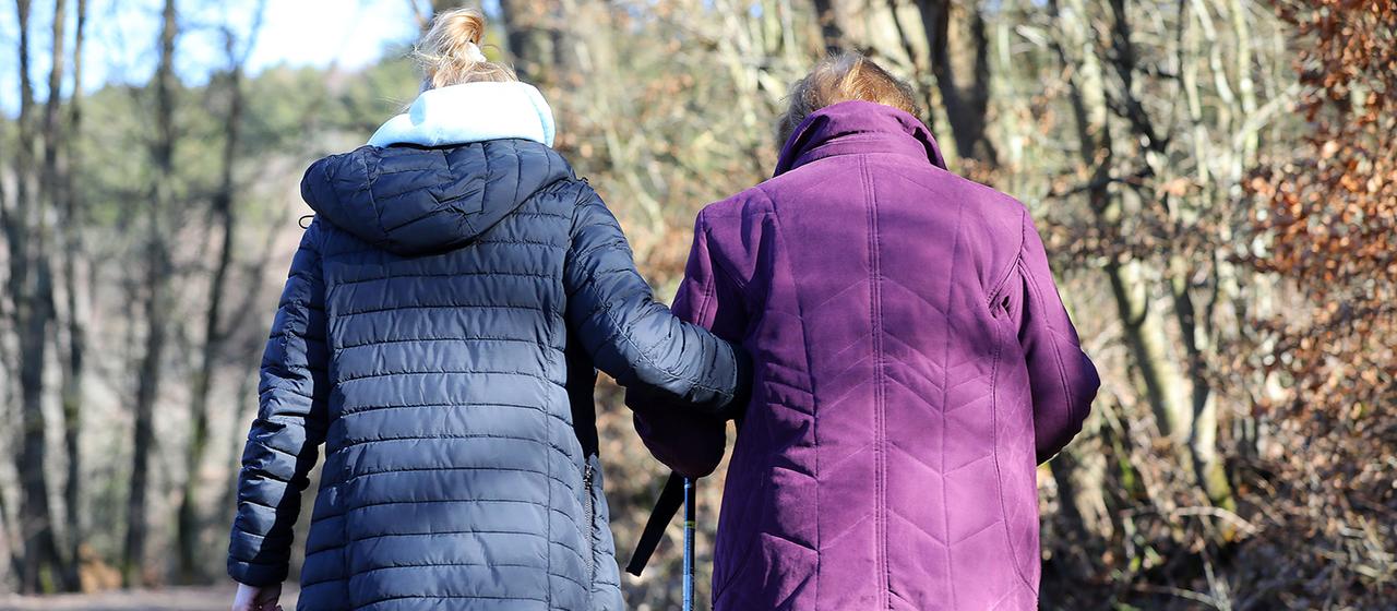 Eine alte Frau mit Stöcken wird von einer jüngeren beim Spazierengehen gestützt.