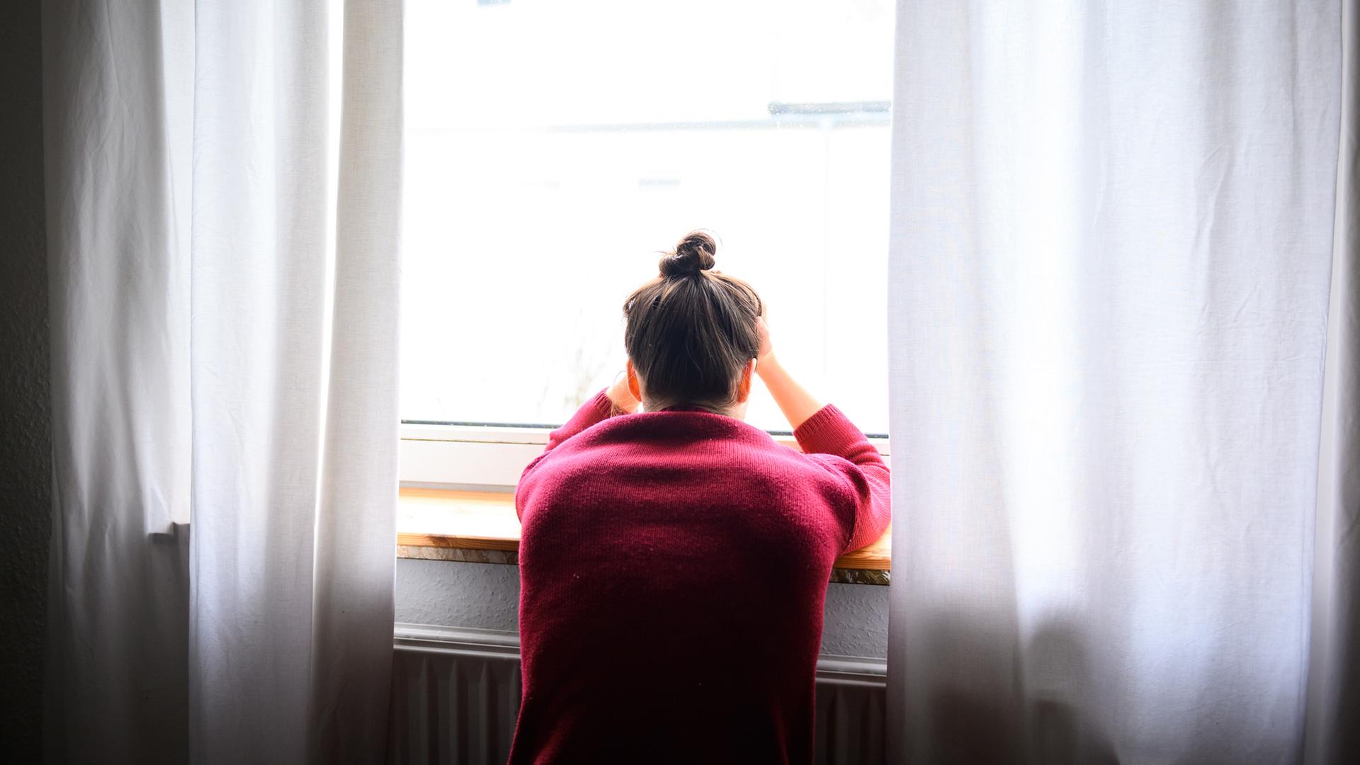 Eine junge Frau sitzt mit Kopf in den Händen am Fenster.