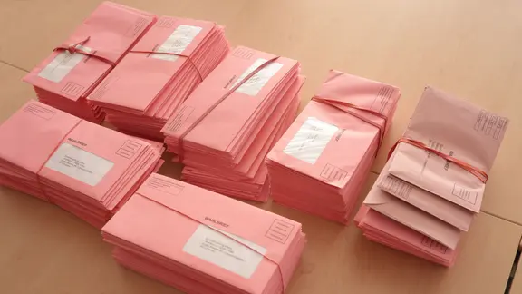 Stapel von roten Wahlbriefen liegen gebündelt auf einem Tisch in einem Briefwahlbüro.