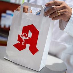 Ein Apotheker legt in einer Apotheke eine Packung mit Tabletten in eine Papiertasche mit Apotheken-Logo.