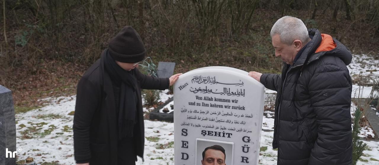 Zwei Personen stehen an einem Grabstein, der mit Blumen geschmückt ist. Auf dem Stein steht in deutscher und arabischer Schrift: "Von Allah kommen wir und zu Ihm kehren wir zurück". Darunter ist ein Foto eines lächelnden Mannes mit der Aufschrift "ŞEHIT SEDAT GÜRBÜZ". Es sind Daten und weiterer Text auf Türkisch vorhanden. Der Boden ist teils mit Schnee bedeckt.