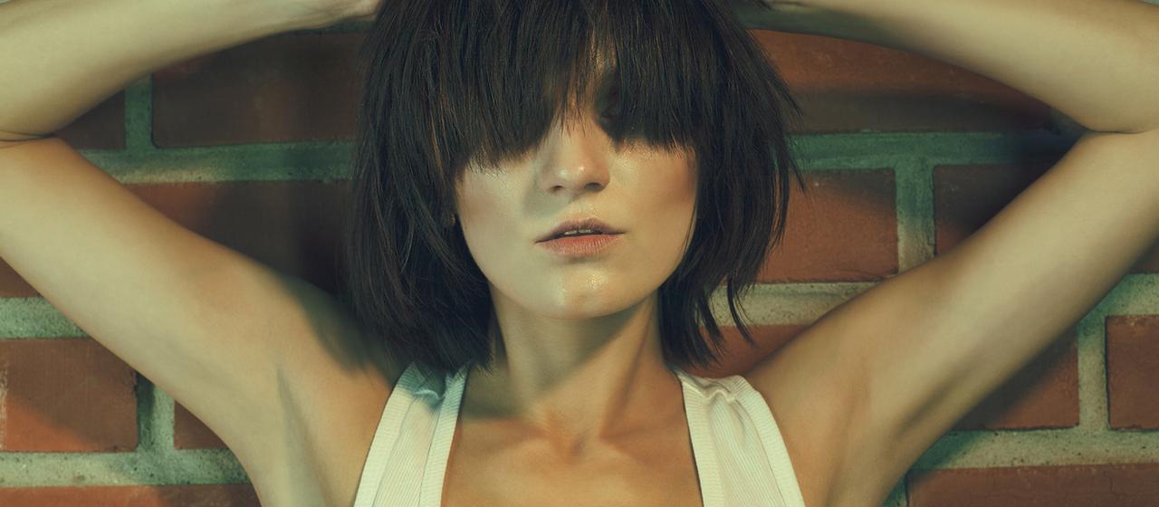 Ein weibliches Model im sogenannten "Heroin-Chic-Stil" steht  vor einer Ziegelmauer.